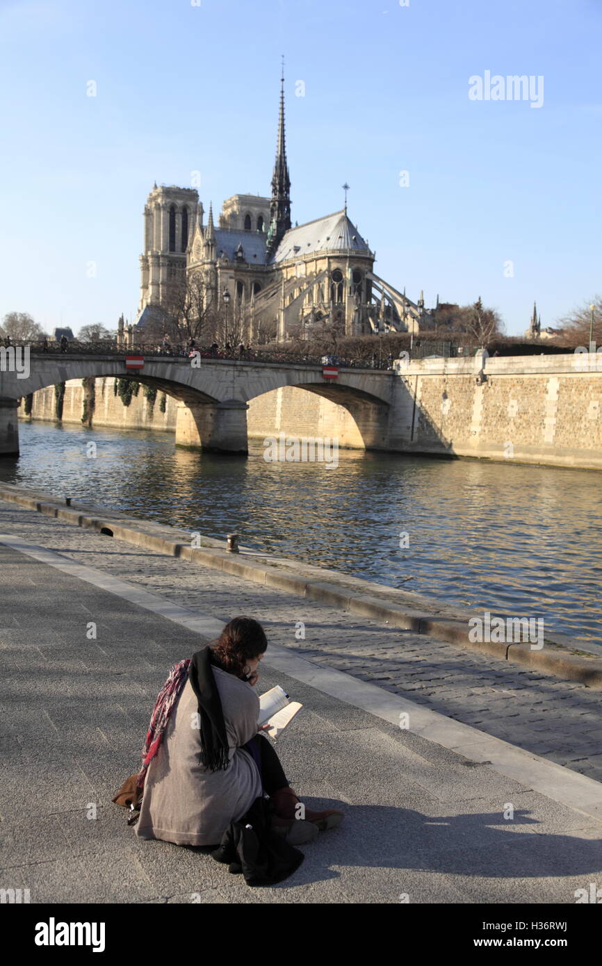 Woman Reading On The Bank Of River Seine With Notre Dame Cathedral