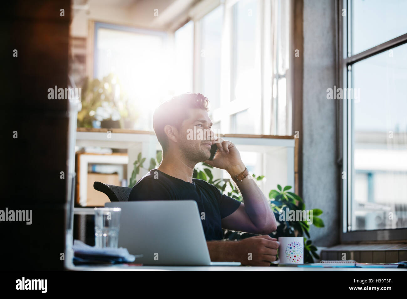 Shot of young businessman sitting at his office and talking on mobile phone. Man working at his desk with laptop making a phone Stock Photo