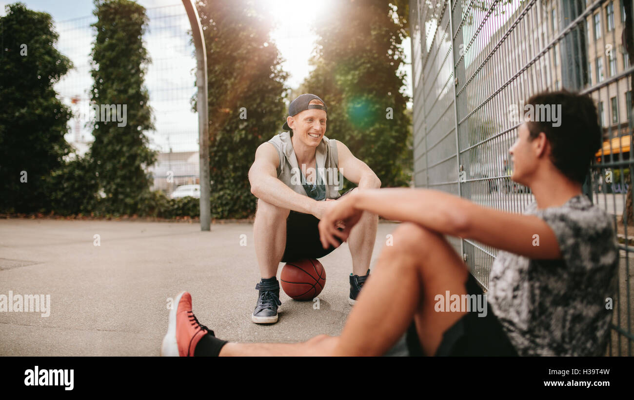 Young friends sitting on basketball court. Streetball players taking rest after playing a game. Two young men relaxing and takin Stock Photo