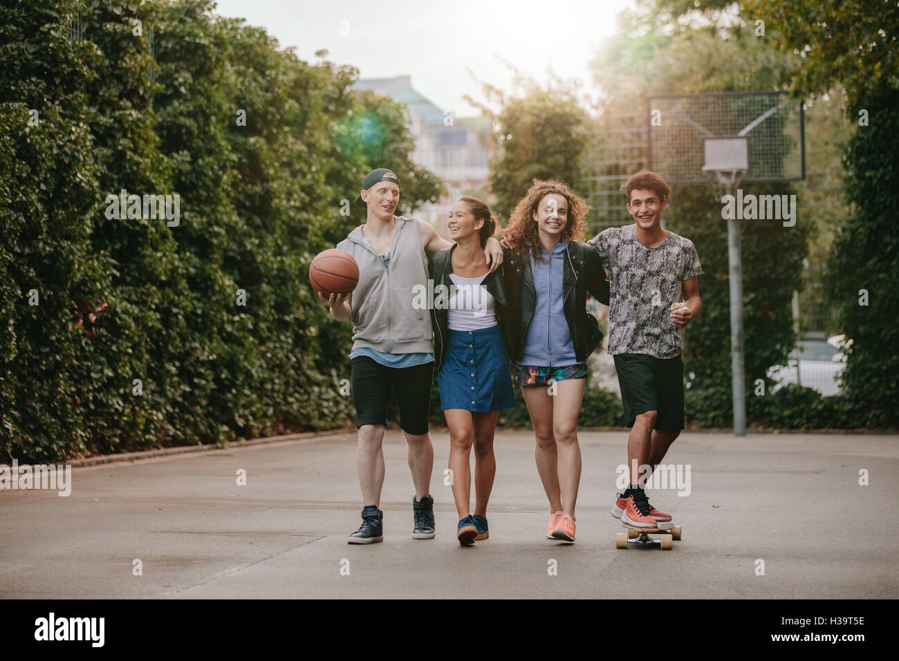 Full length shot of happy teenage friends enjoying on basketball court. Young men and women walking with skateboard and basketba Stock Photo