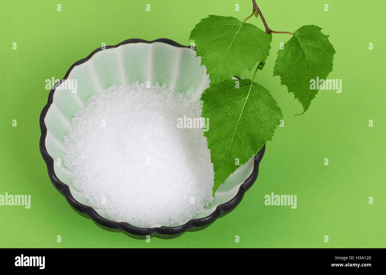 Xylitol birch sugar in white porcelain bowl with birch leaves over green. White granulated sugar alcohol, substitute sweetener. Stock Photo