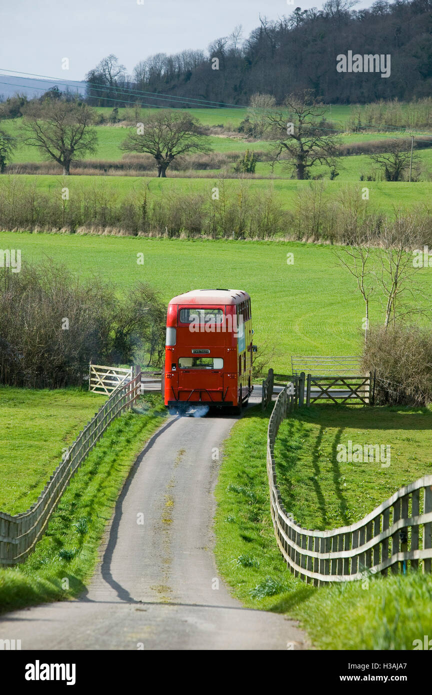 An old double decker red London bus driving through the Biritish  countryside on a journey taking it past rolling hills,fieldds adn trees. In  a scene remeniscent of the famous Summer Holiday film