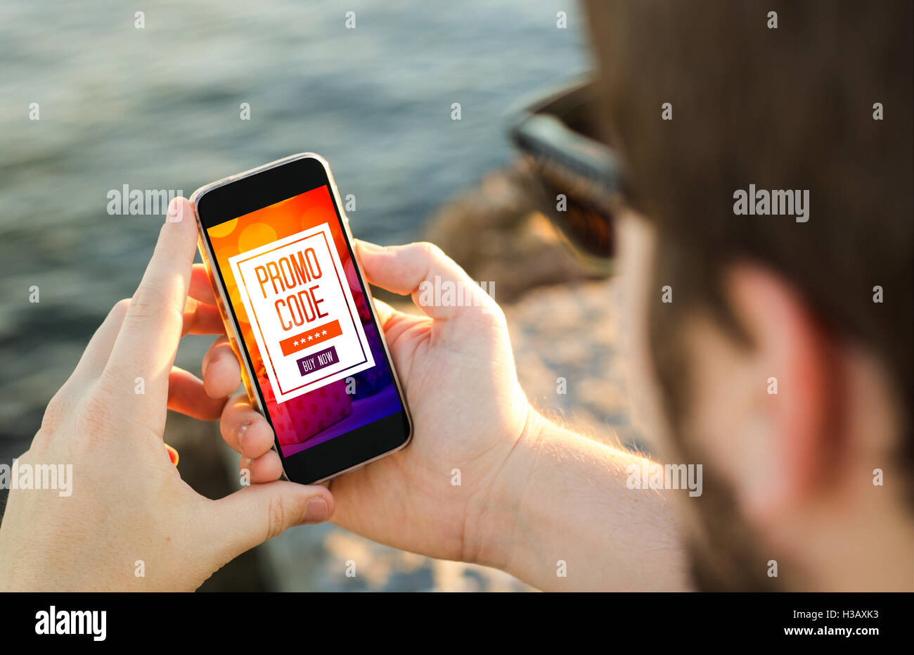 M-commerce concept: man on the coast holding his phone and using a promo code. All screen graphics are made up. Stock Photo