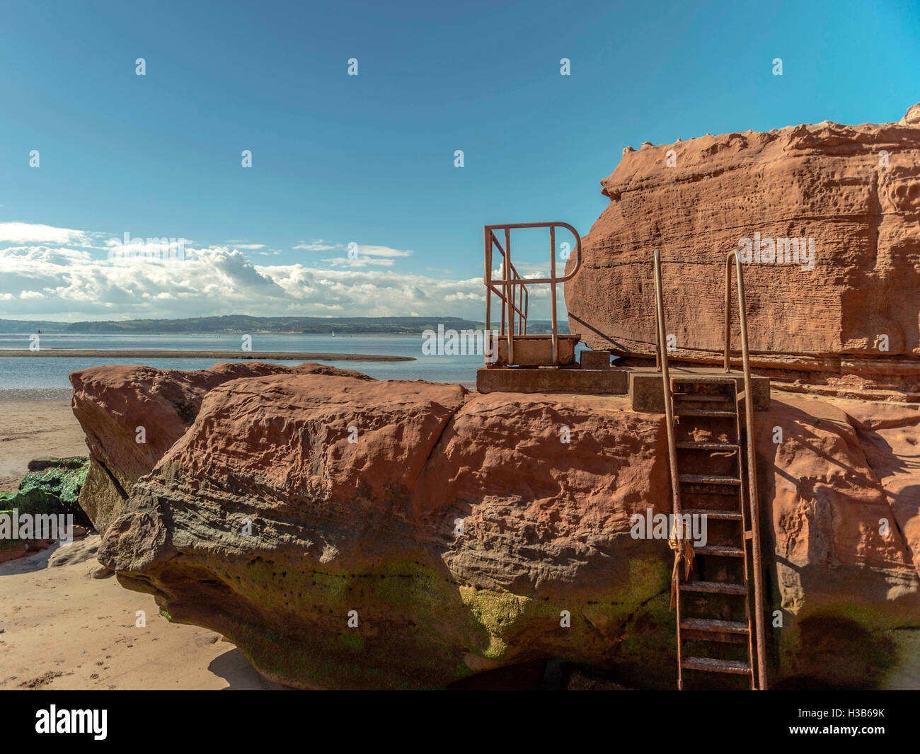 Rustic rock formations visible at low tide and rusting metal gangways at Orcome Point near the seaside town of Exmouth, Devon. Stock Photo