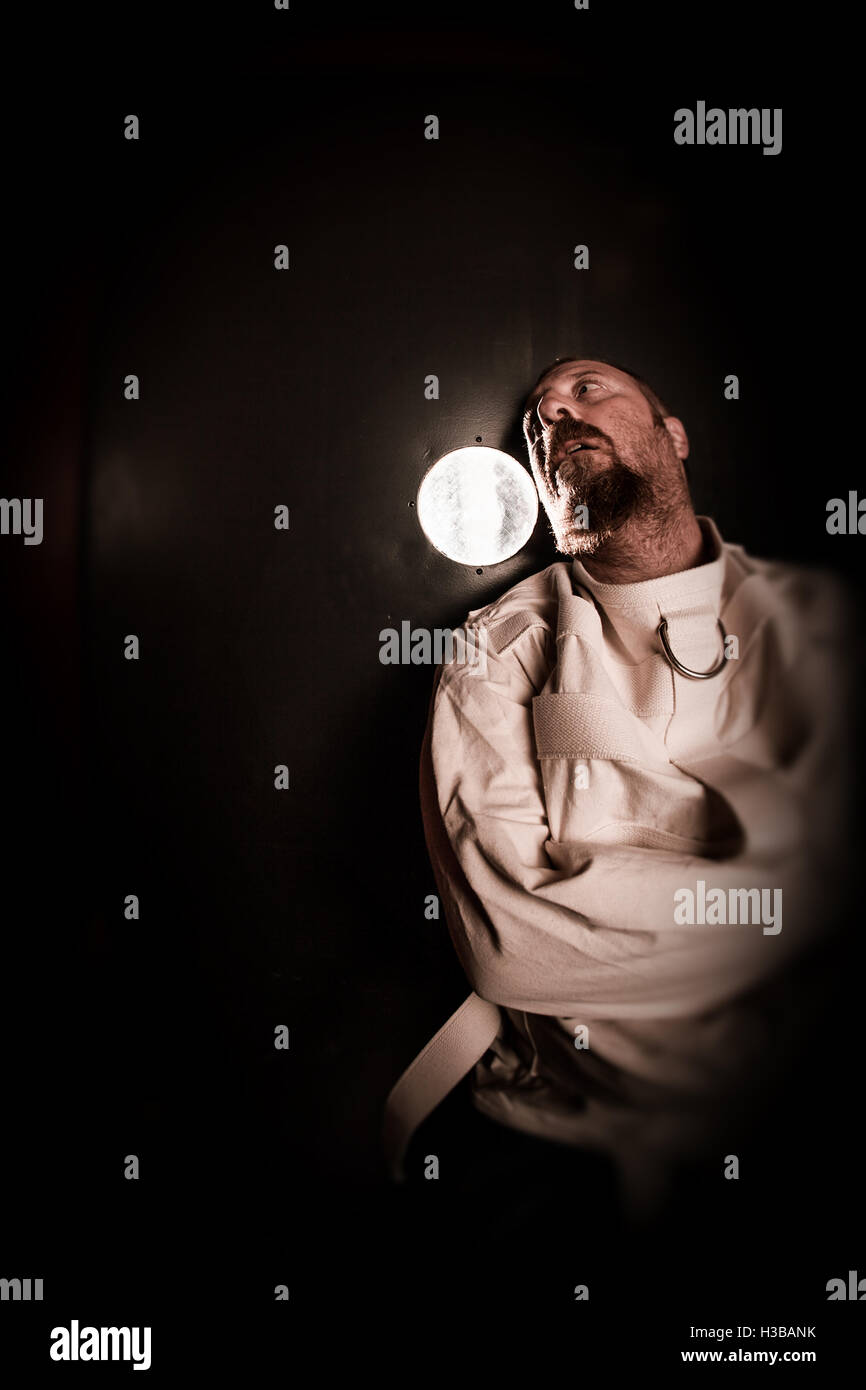 Photo of an insane man in his forties wearing a straitjacket standing in a cell of an asylum with the light from the hallway. Stock Photo