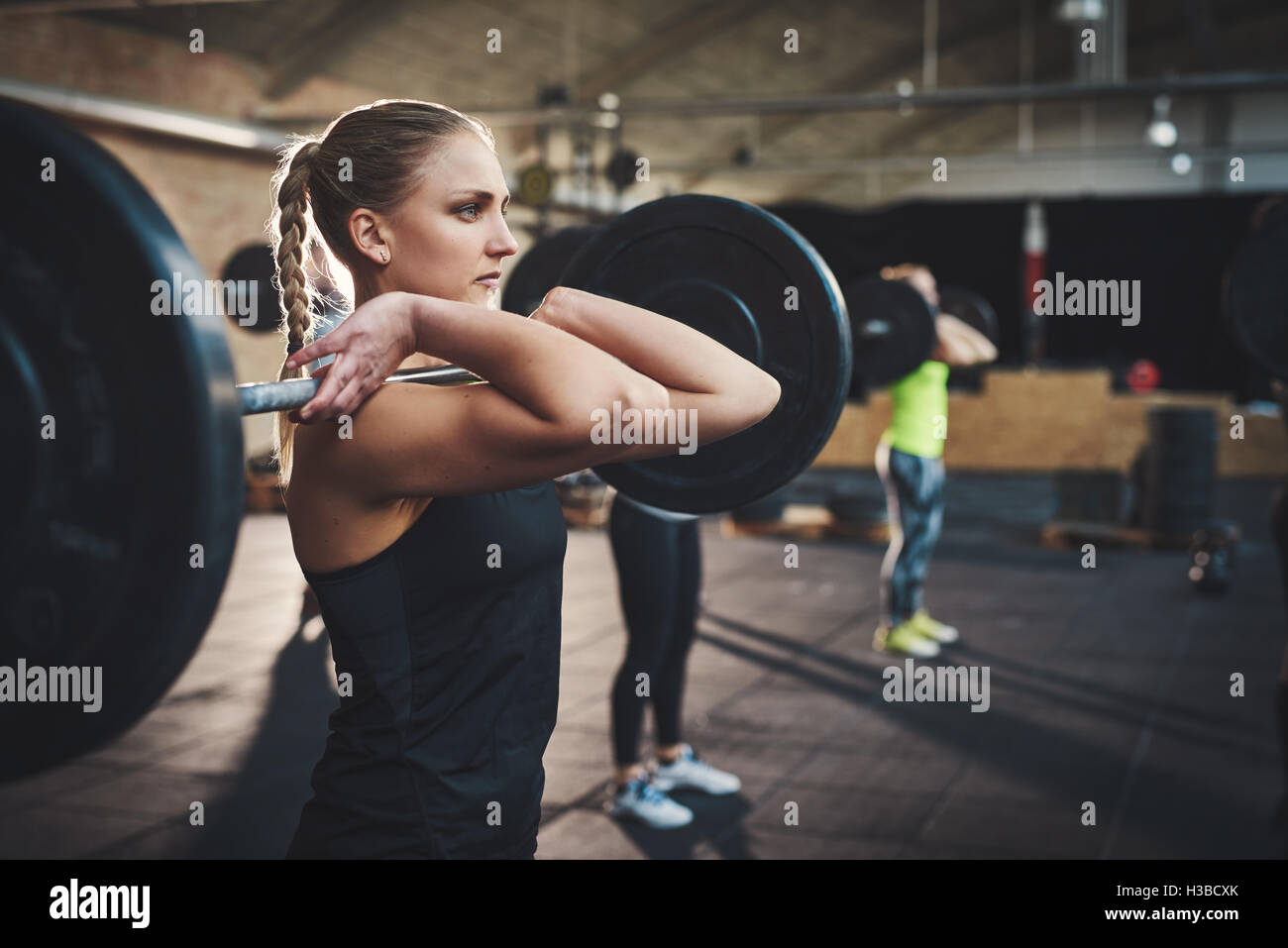 Tough woman with thick muscular arms pulling up large barbell in fitness training class with other adults and black floor mats i Stock Photo