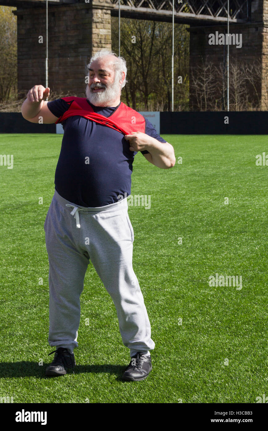 Overweight middle-aged man struggling  to put on a bib shirt ready to take part in a walking  football session. Stock Photo