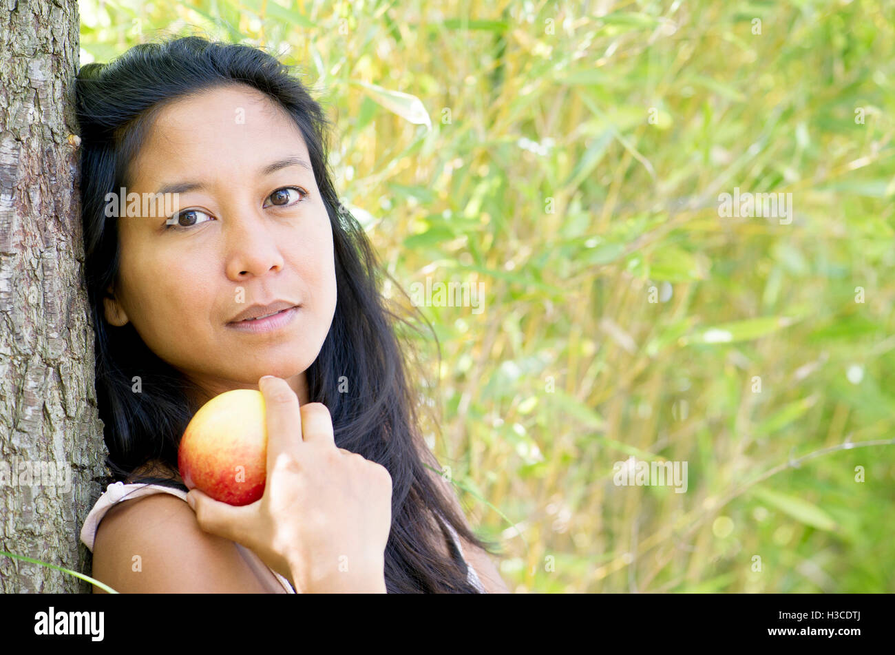 Woman leaning against tree trunk with apple in hand, portrait Stock Photo
