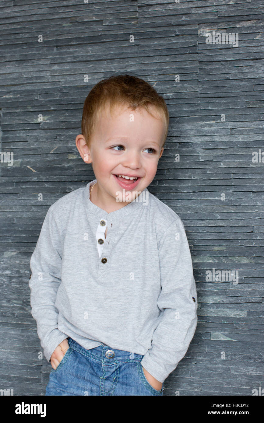 Little boy leaning against wall with hands in pockets, smiling cheerfully Stock Photo