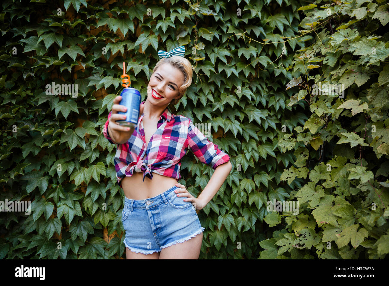 Happy beautiful pin up girl giving you a soda can and winking in the garden Stock Photo
