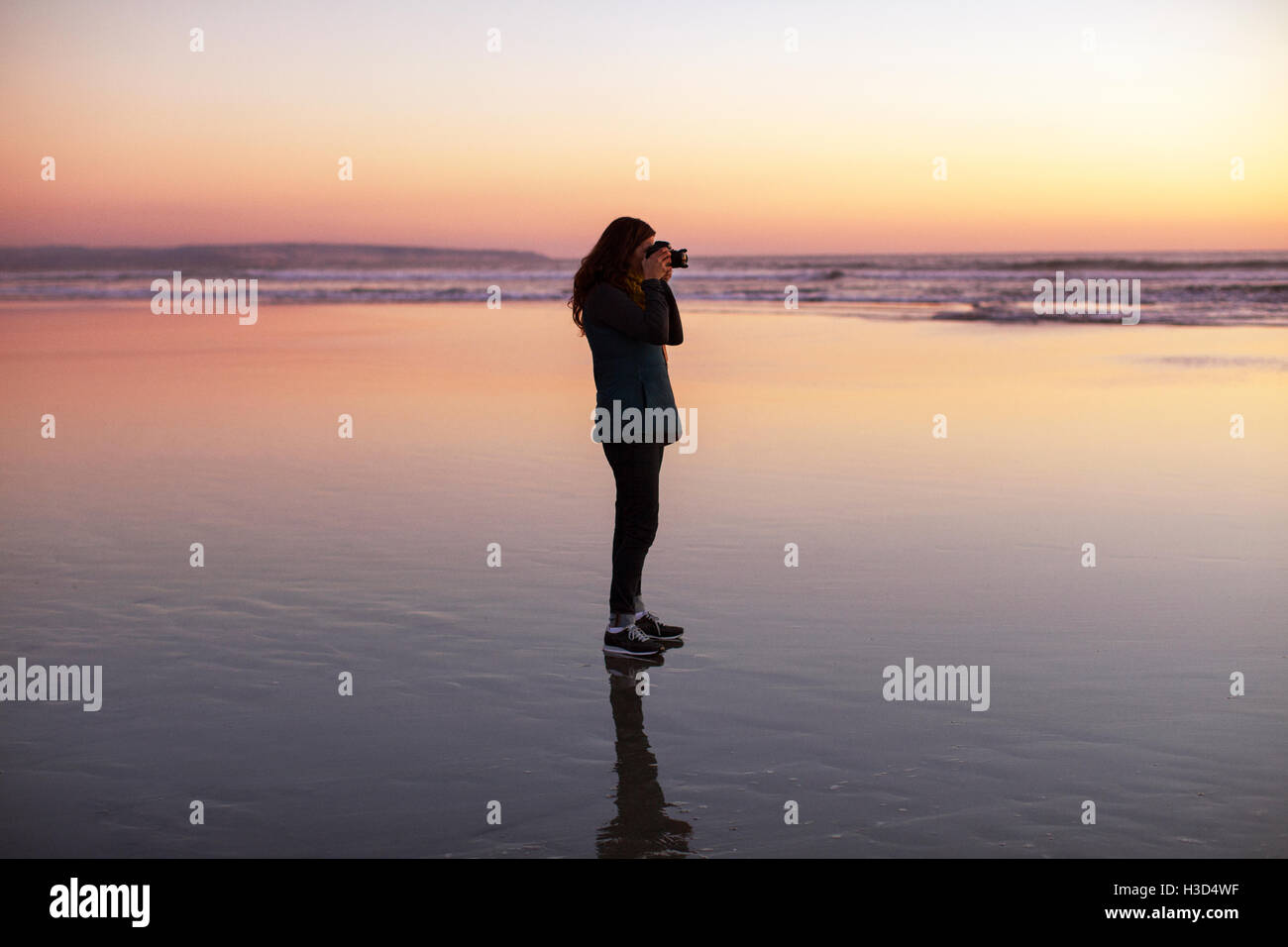 Side view of woman photographing while standing at beach during sunset Stock Photo