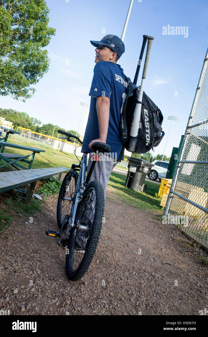 Young teen baseball player in uniform carrying two bats with his bicycle as mode of transportation. St Paul Minnesota MN USA Stock Photo