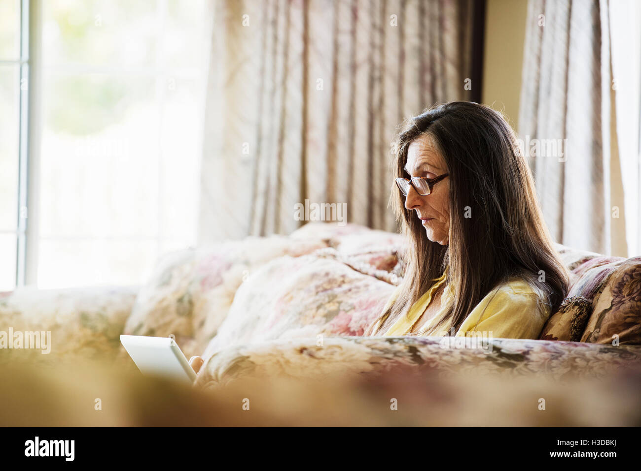 Senior woman with long brown hair sitting on a sofa, using a laptop computer, wearing reading glasses. Stock Photo