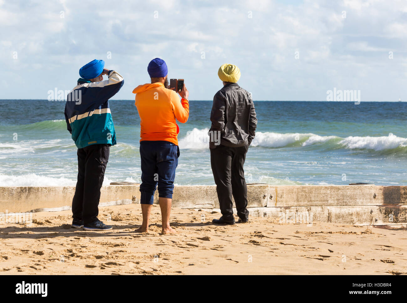 Three male tourists standing on Bournemouth beach taking a photo on mobile phone on a windy day in October Stock Photo