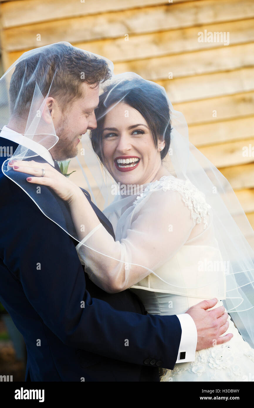 A bride and groom, a young woman laughing and draping her veil over her husband's head. Stock Photo