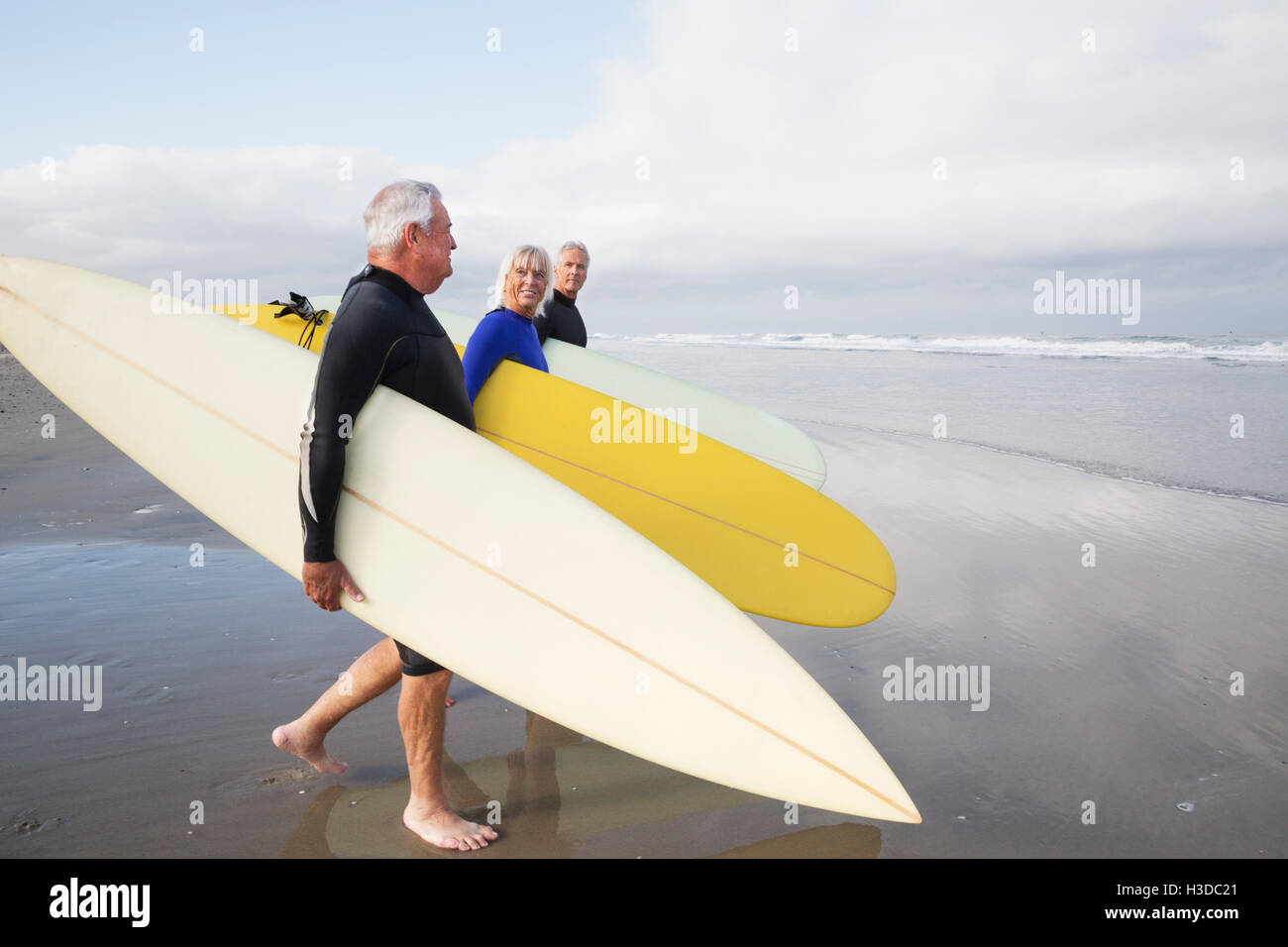 Senior woman and two senior men on a beach, wearing wetsuits and carrying surfboards. Stock Photo
