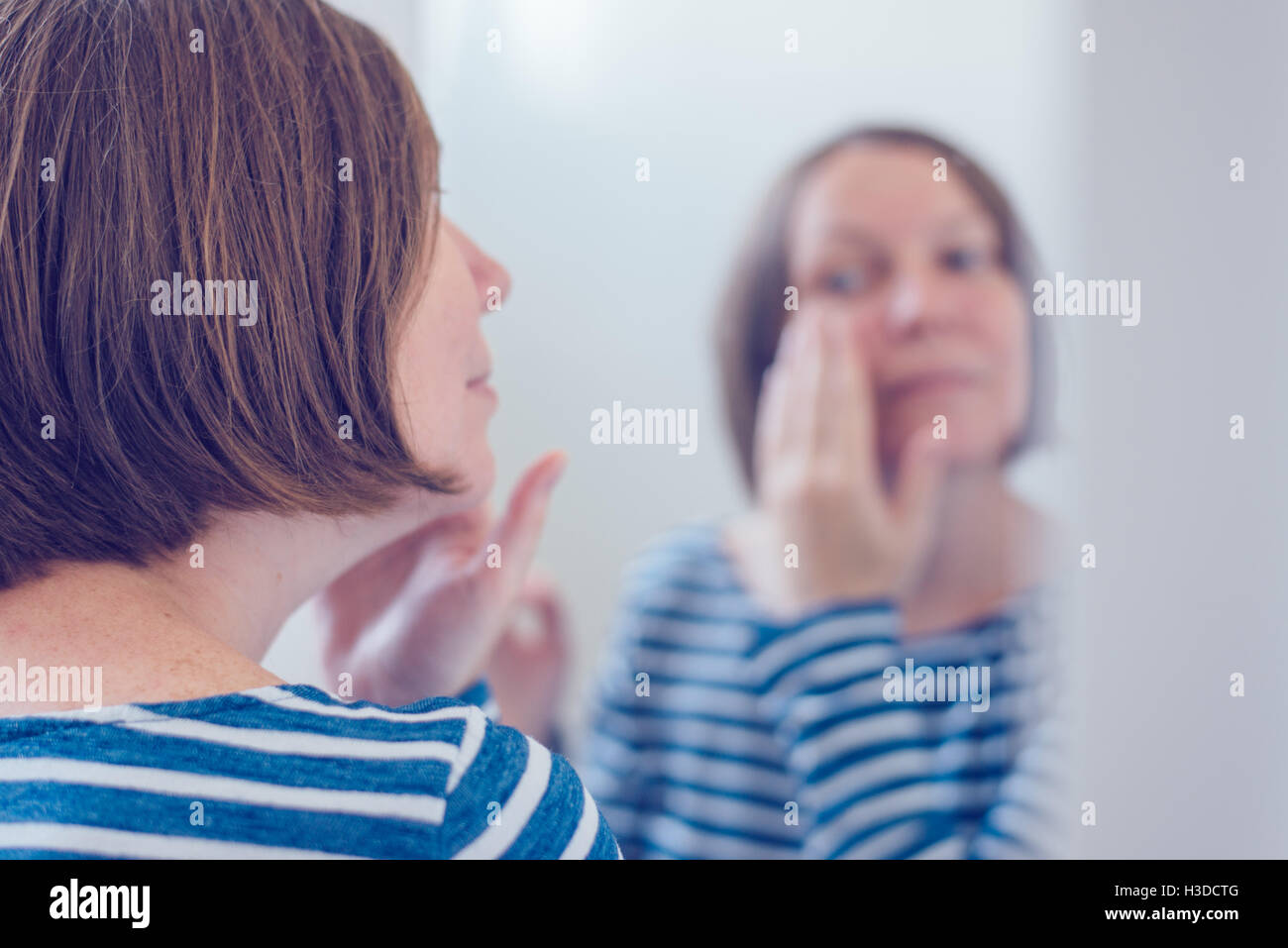 Young adult woman applying anti-aging cream in front of the bathroom mirror, selective focus Stock Photo
