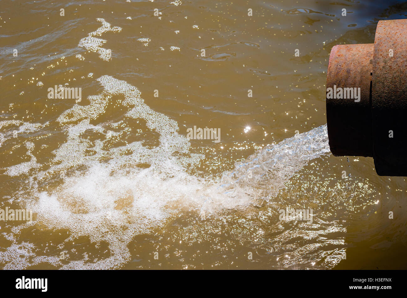Water flowing from rusted metal culvert drain pipe opening and splashing into foamy brown water. Stock Photo
