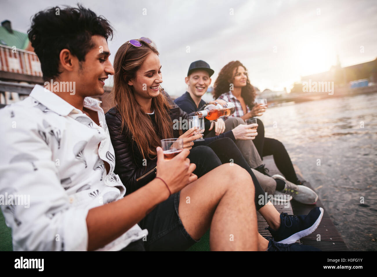 Shot of group of people sitting outdoors on jetty and having drinks. Friends hanging out on pier over lake in the city. Stock Photo