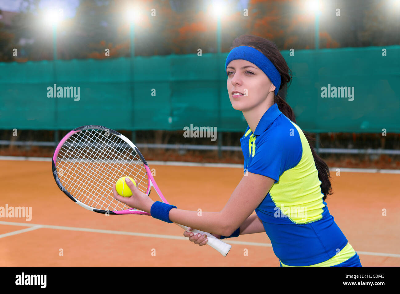 Beautiful young female tennis player serving wearing a sportswear during a tennis match on clay court  background with lights outdoor in evening. Stock Photo