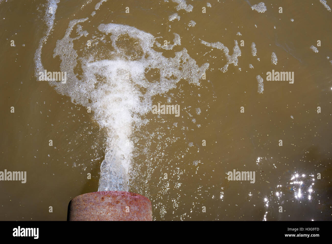 Water flowing from rusted metal culvert drain pipe opening and splashing into foamy brown water, viewed from above. Stock Photo