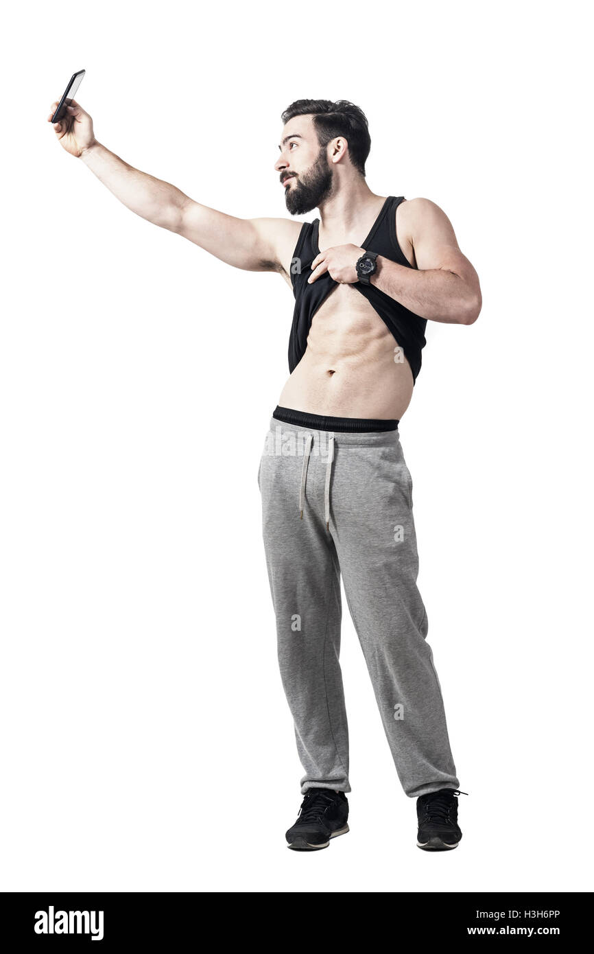 Muscular fit man showing abs muscles while taking selfie with smart phone. Full body length portrait isolated on white Stock Photo