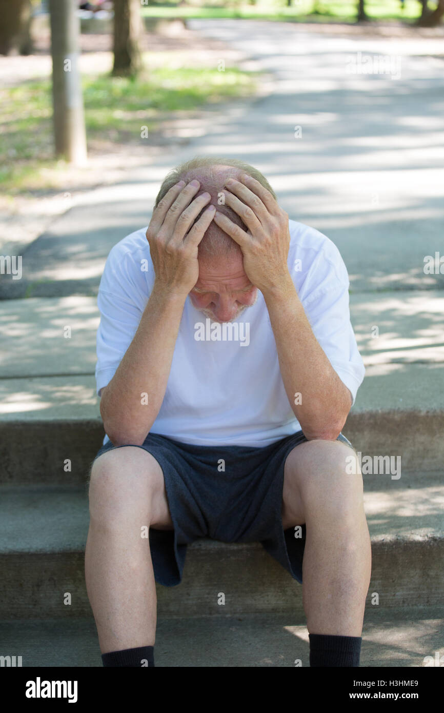 Closeup portrait, senior mature man in black shorts and white shirt, sitting down outside on steps, hands on head in defeat Stock Photo