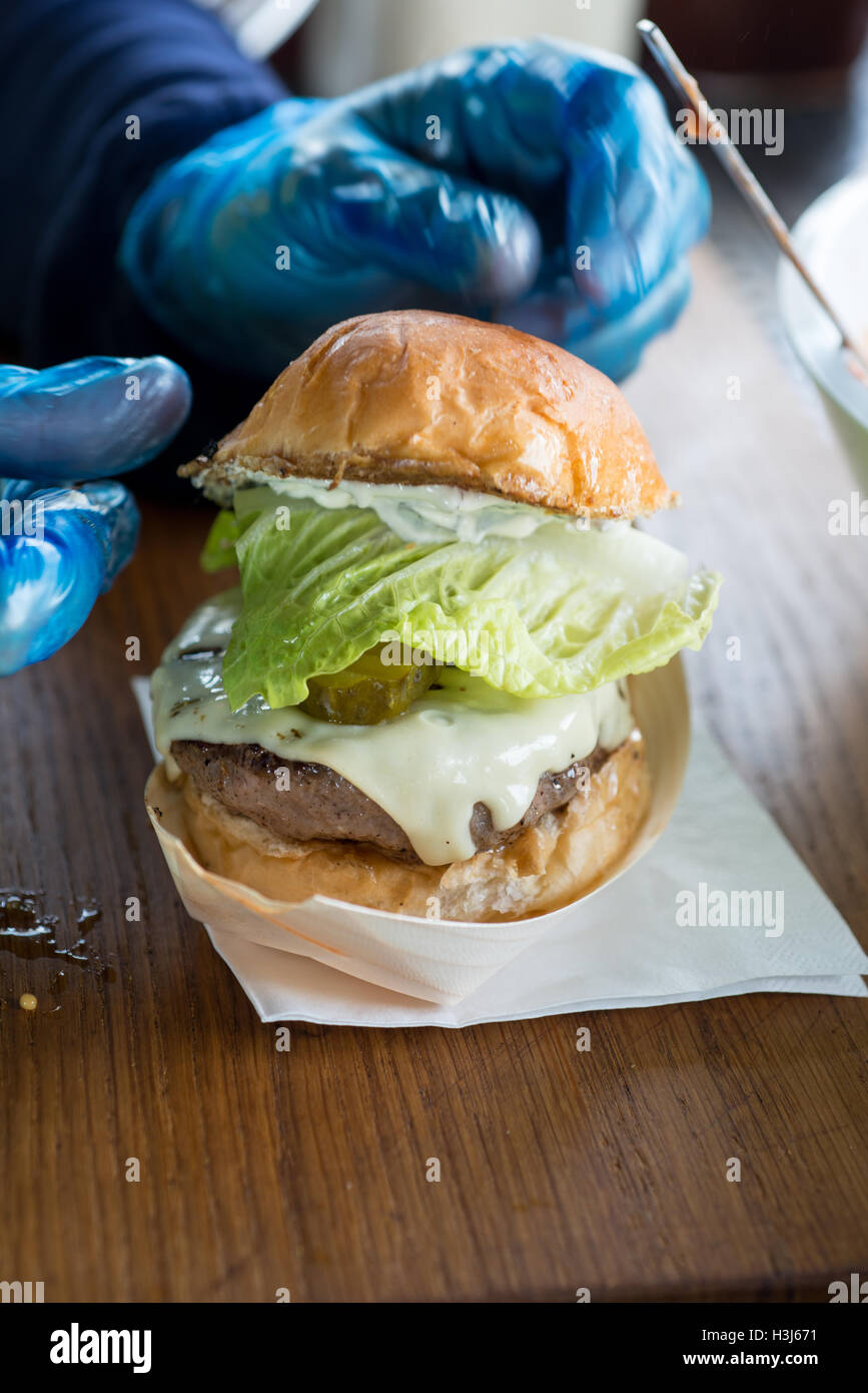 cheeseburger with lettuce and gherkins in a bread bun in a reflective food tray on a napkin on a wooden surface with gloved hand Stock Photo