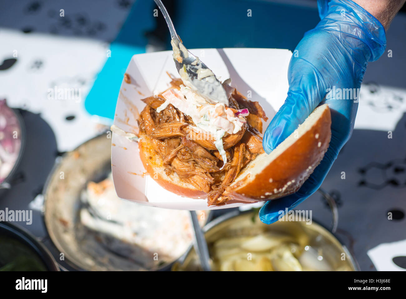 white bread bun in a white food tray being topped with toppings such as pulled pork and coleslaw with gloved hands holding tongs Stock Photo