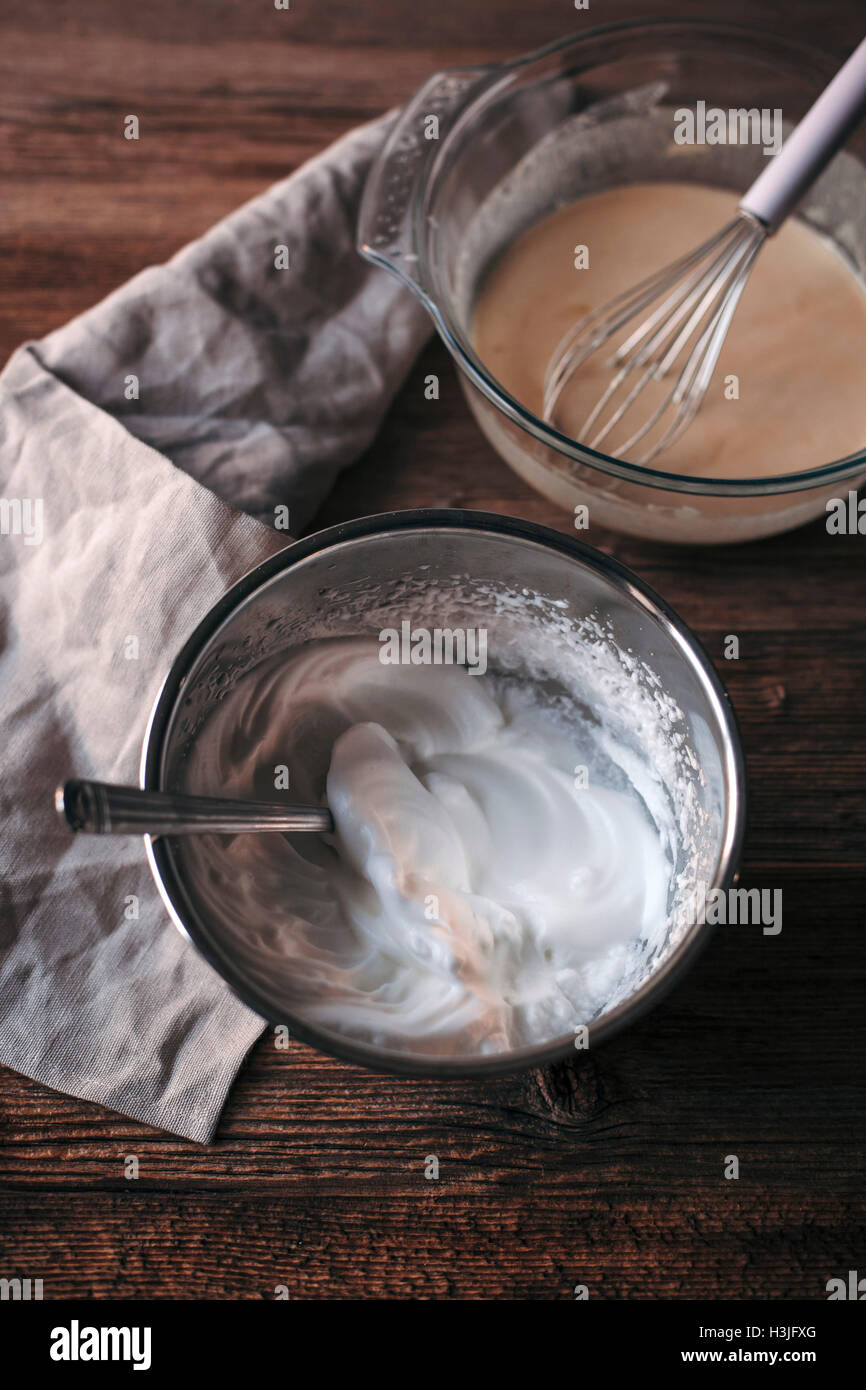 Eggs, sugar, milk, flour batter in a glass bowl and whipped eggs whites in a mixing bowl on a wooden table Stock Photo