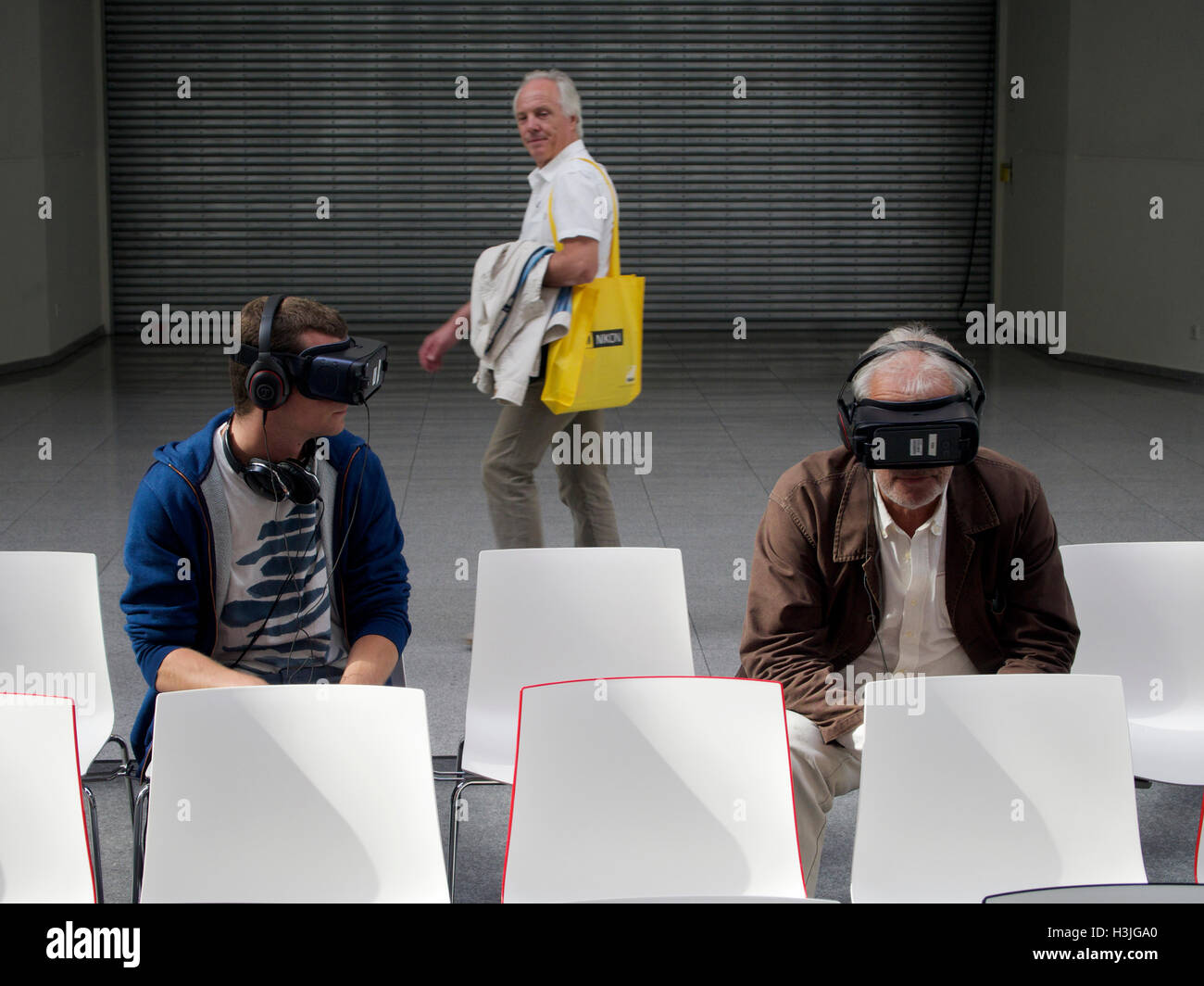 Two men wearing VR glasses in boring and sterile environment, with third man passing looking at them. Cologne, Germany Stock Photo