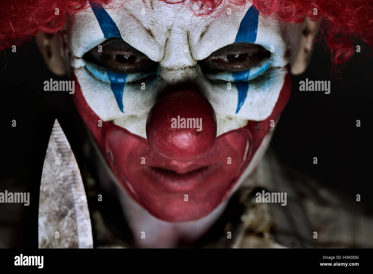 closeup of a scary evil clown wearing a ragged shirt, holding a big knife next to his face Stock Photo