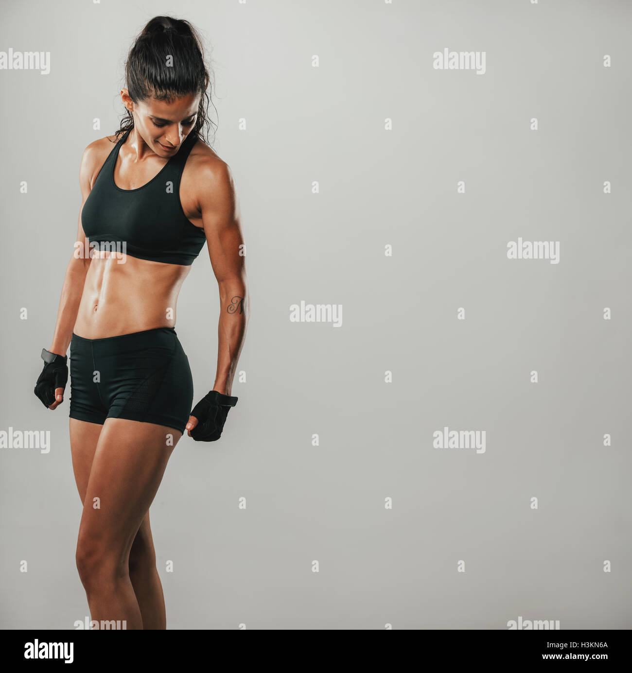 Muscular woman in black sports shorts and top looks down while standing in a grey room Stock Photo