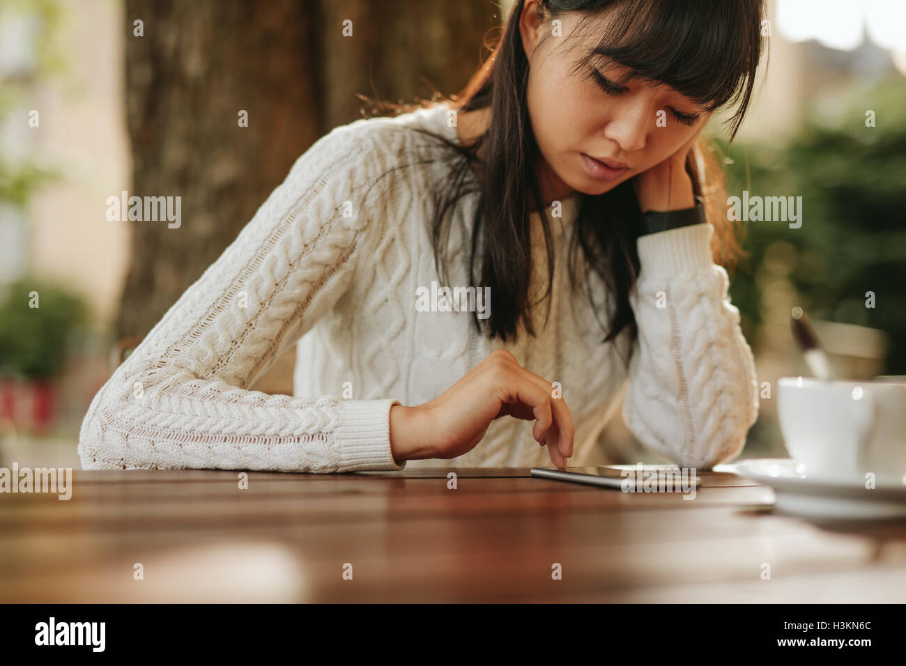 Young chinese woman looking at her cellphone while sitting at outdoor cafe. Asian female model using mobile phone at coffeeshop. Stock Photo