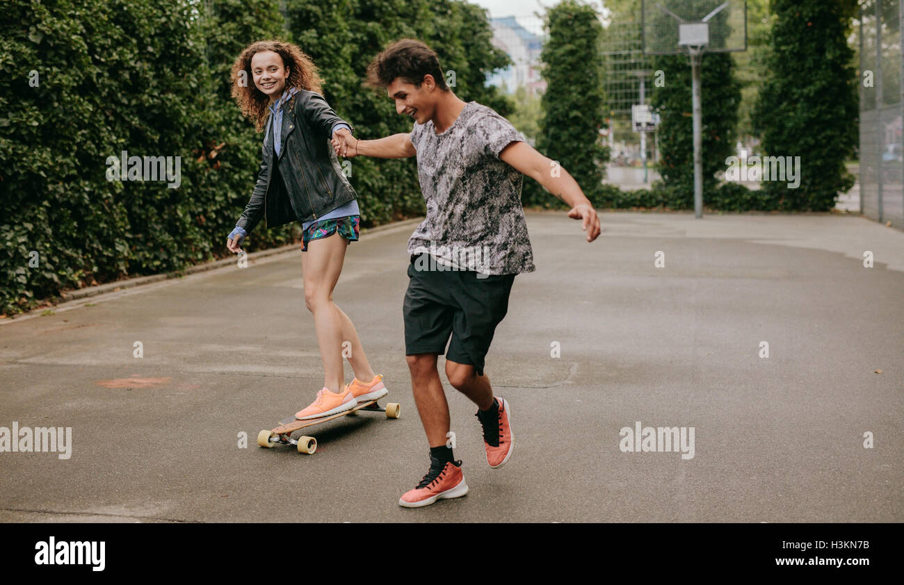 Young woman standing on a longboard with support from her boyfriend. Woman skating on a basketball court with friend. Stock Photo