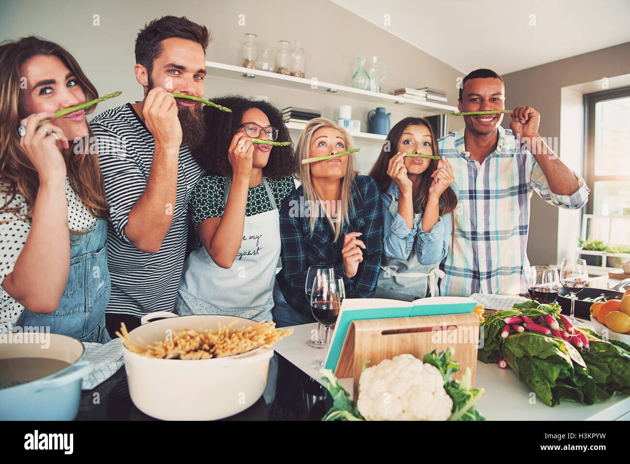 Group of six diverse silly adults sniffing asparagus stalks in kitchen. Bowl of pasta and vegetables are on the table. Stock Photo
