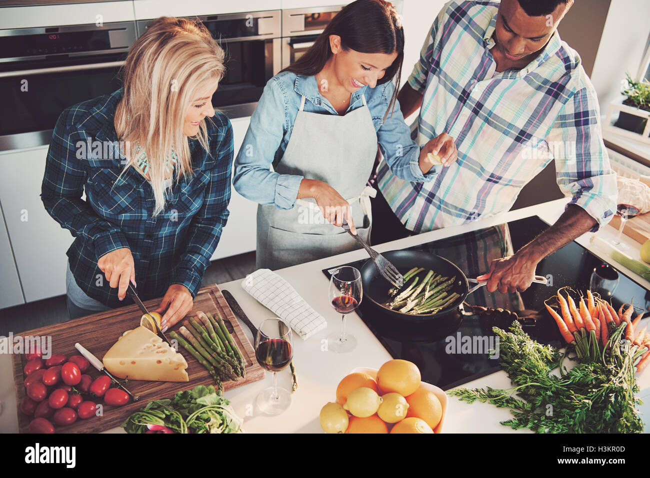 High angle view of trio cooking a meal with tomatoes, cheese, asparagus, fruit and other ingredients on kitchen counter and stov Stock Photo