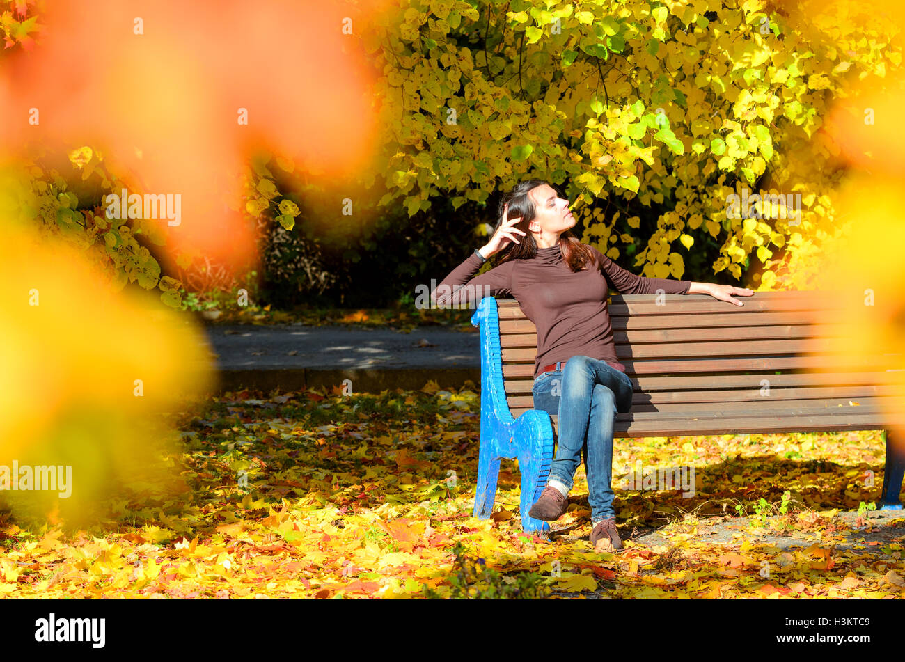 Attractive young woman sitting on brown wooden bench with hands behind head in beautiful park. She half-closed her eyes. Golden autumn foliage around. Girl wears blue jeans and turtleneck sweater Stock Photo