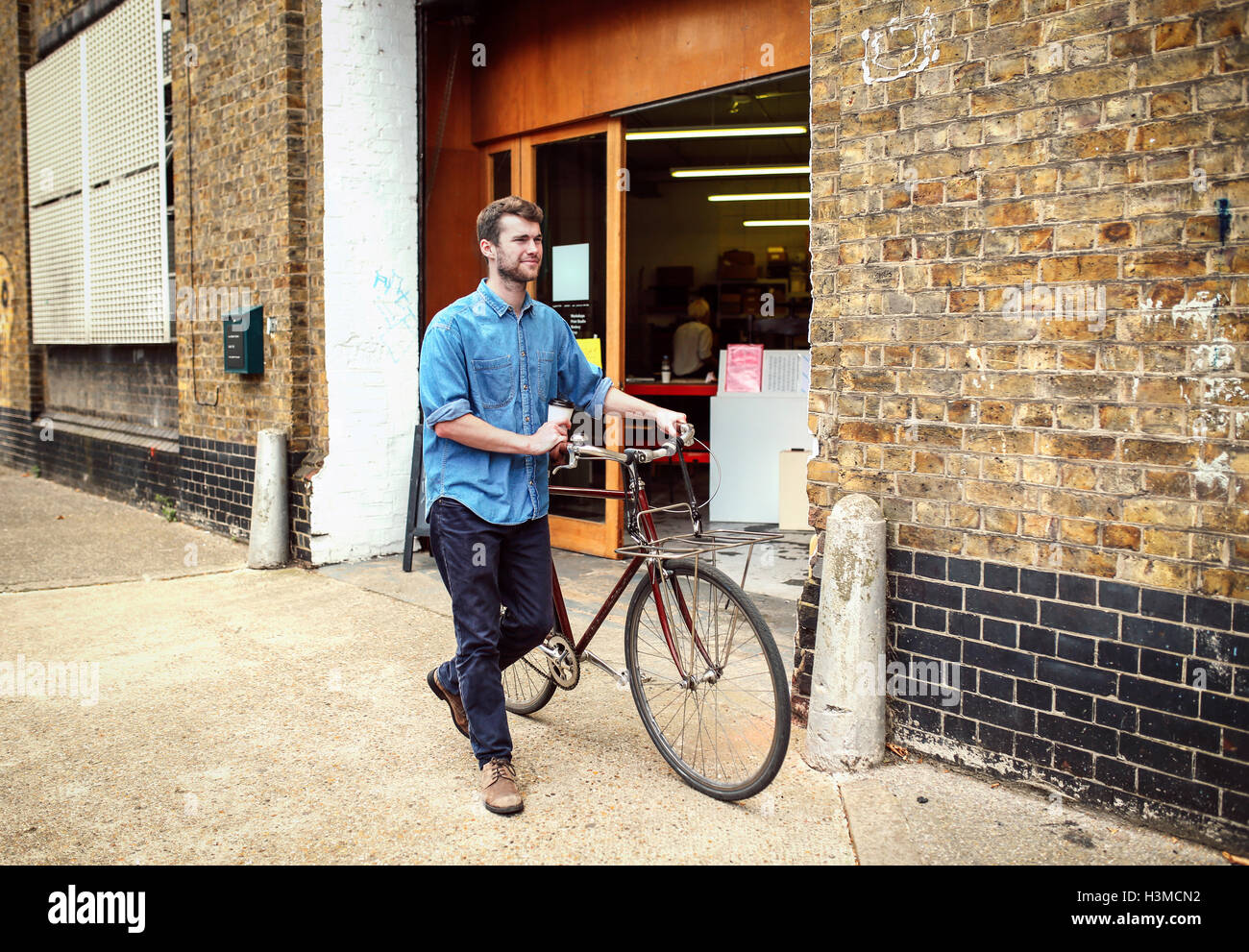 Young man carrying coffee and wheeling pushbike past workshop Stock Photo