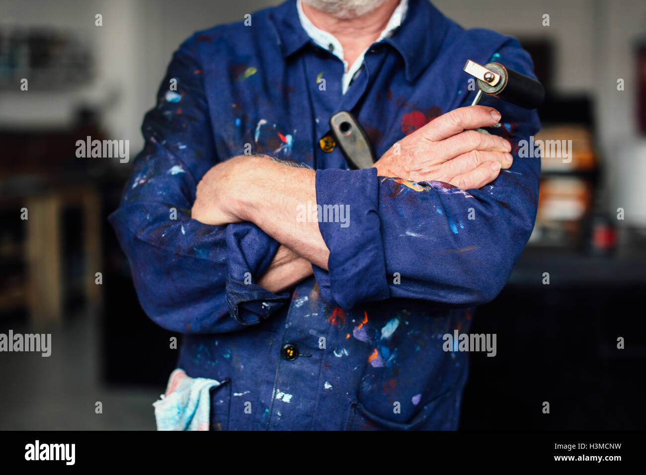 Senior craftsman with arms folded in print workshop, holding ink roller and wearing paint-splattered overalls, mid section Stock Photo