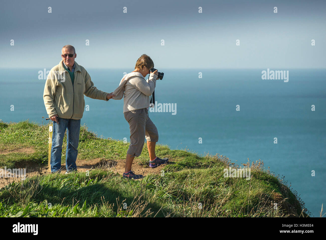 A man holds on to his wife’s clothing as she stand on the edge of a cliff to take a photograph. Stock Photo