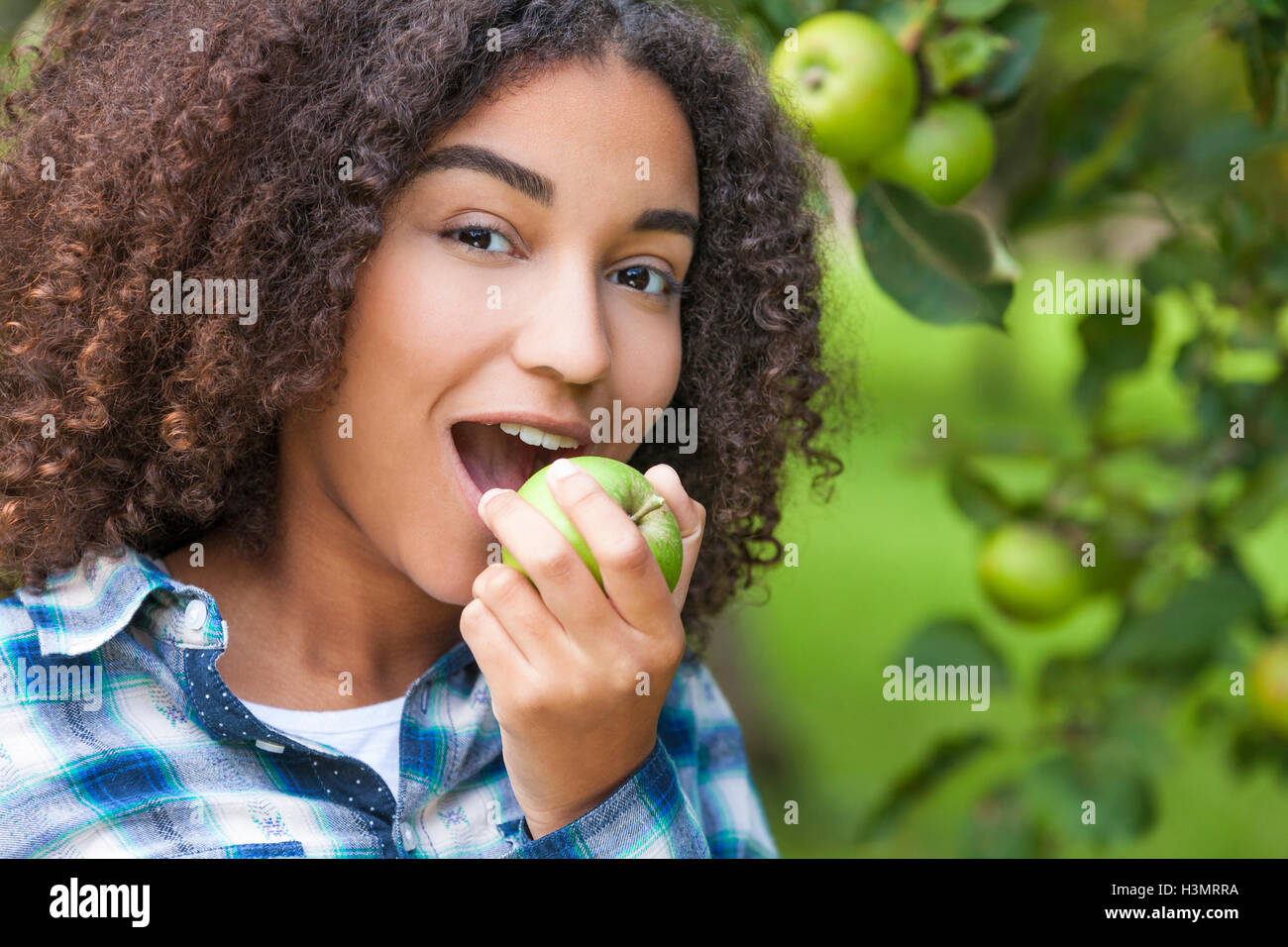 Outdoor portrait of beautiful happy mixed race African American girl teenager female child eating an organic green apple Stock Photo