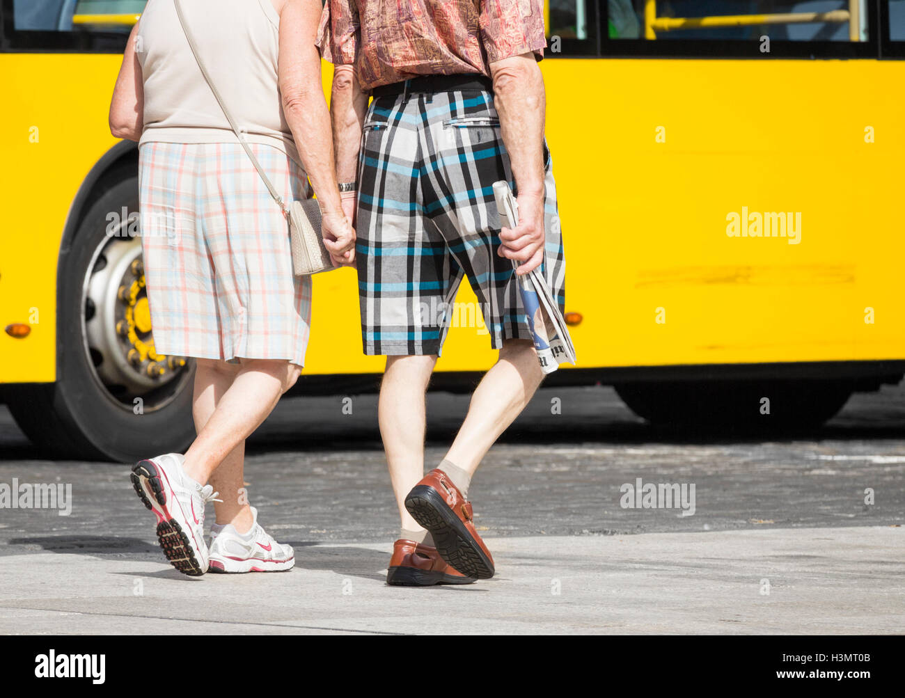 Elderly couple in checked shorts holding hands on holiday in Spain. Man holding copy of English newspaper. Stock Photo