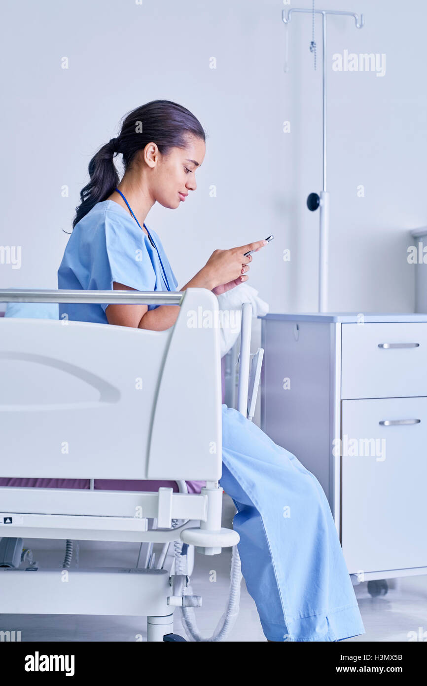Female medic sitting on hospital bed reading smartphone text messages Stock Photo