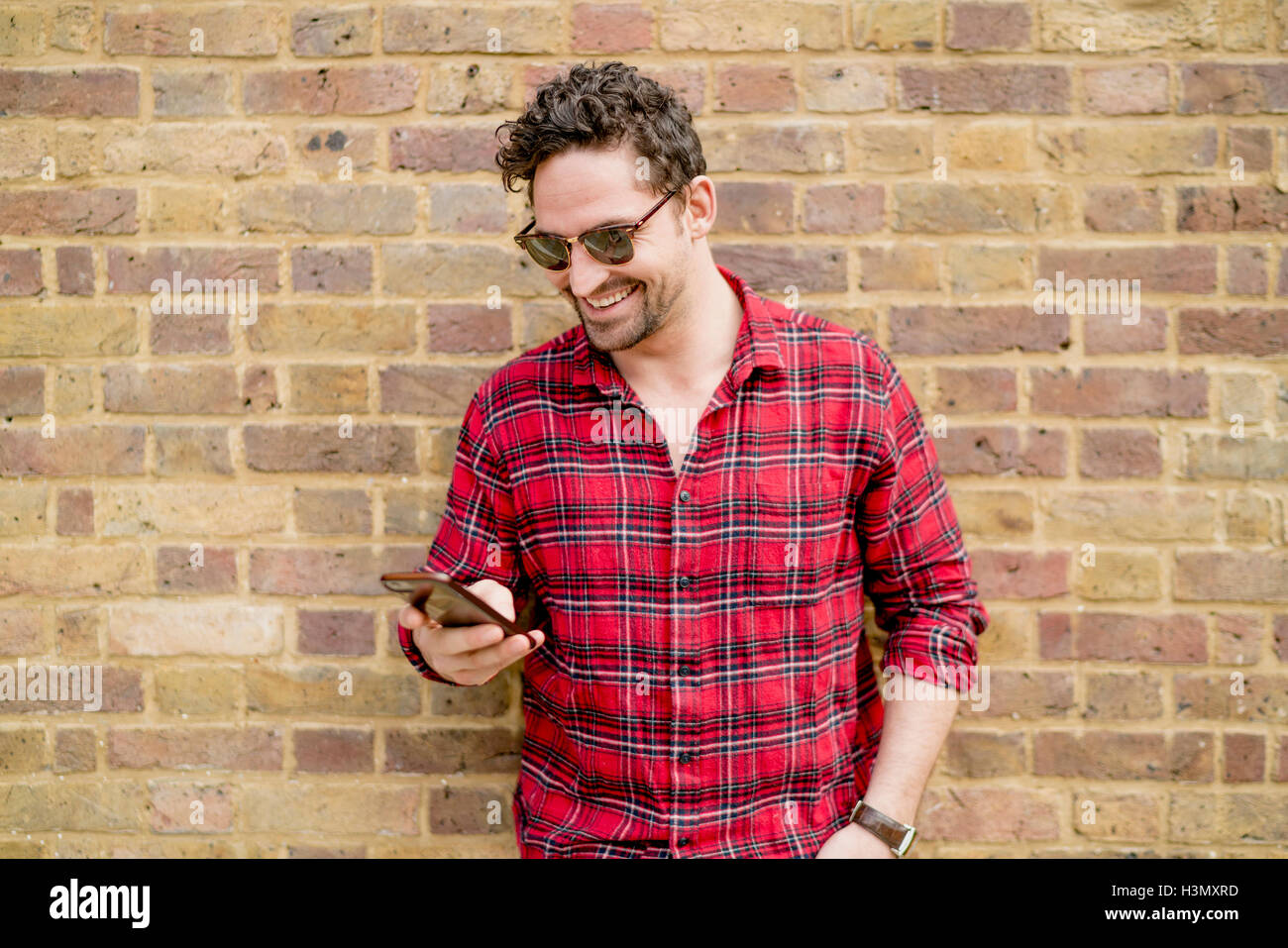 Young man leaning against brick wall reading smartphone texts Stock Photo