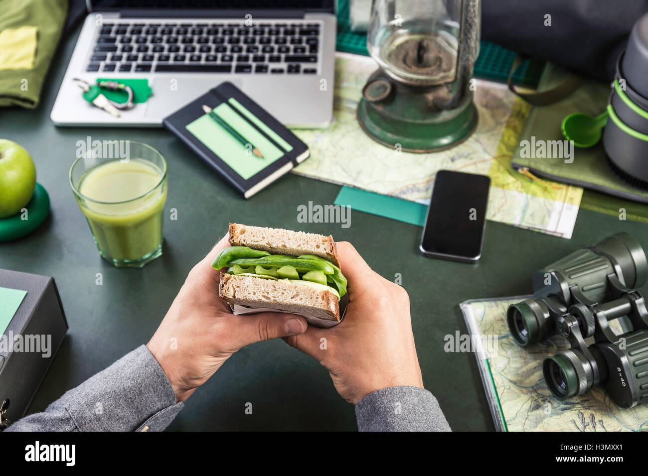 Mans hand's holding sandwich at table with hiking equipment and laptop Stock Photo