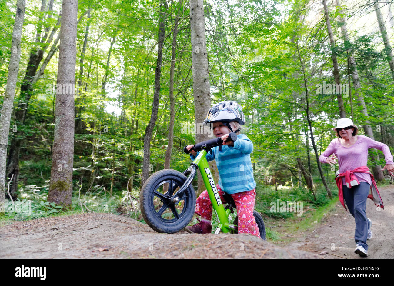 A 2 yr old girl on a balance bike with her grandmother fearfully chasing. Taken on Leapfrog pump track at Kingdom Trails Vermont Stock Photo