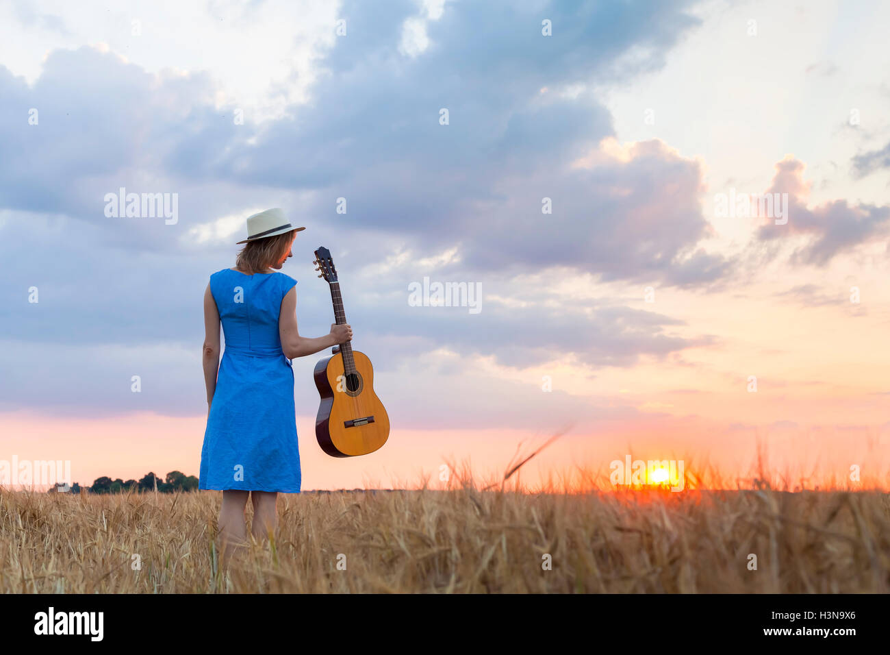 Young pretty woman walking in wheat field at sunset with acoustic guitar in hand to find inspiration for country songs Stock Photo