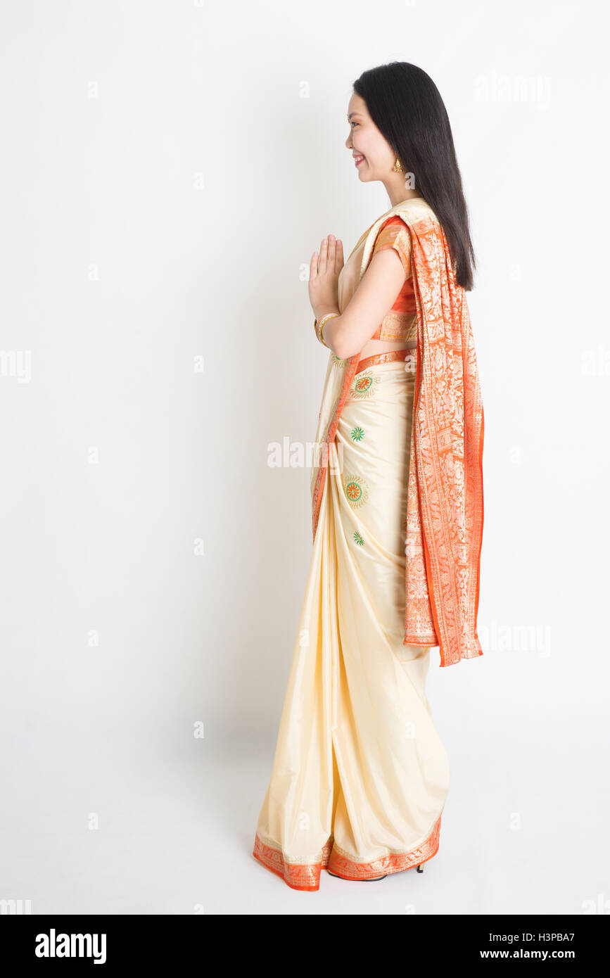 Full length side view mixed race Indian Chinese female with sari dress in greeting gesture, standing on plain background. Stock Photo