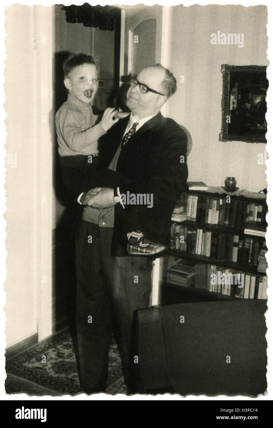 GERMANY - CIRCA 1970: Bald businessman wearing glasses and a business suit is holding a little boy in shorts. He is standing in the living room Stock Photo